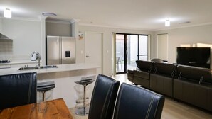 Eine moderne Küche mit weißen Schränken, einem Edelstahlkühlschrank und einer Kaffeemaschine. Ein Essbereich mit einem Holztisch und schwarzen Ledersesseln ist ebenfalls vorhanden. Im Wohnbereich sind eine Couch und ein Fernseher zu sehen.