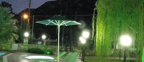 A patio area with lounge chairs and a green umbrella, a pool, and a weeping willow tree.