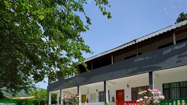 An outdoor dining area with a table set for a meal, surrounded by greenery and a building with a red door. - ONES PANSİYON (Kumluca)