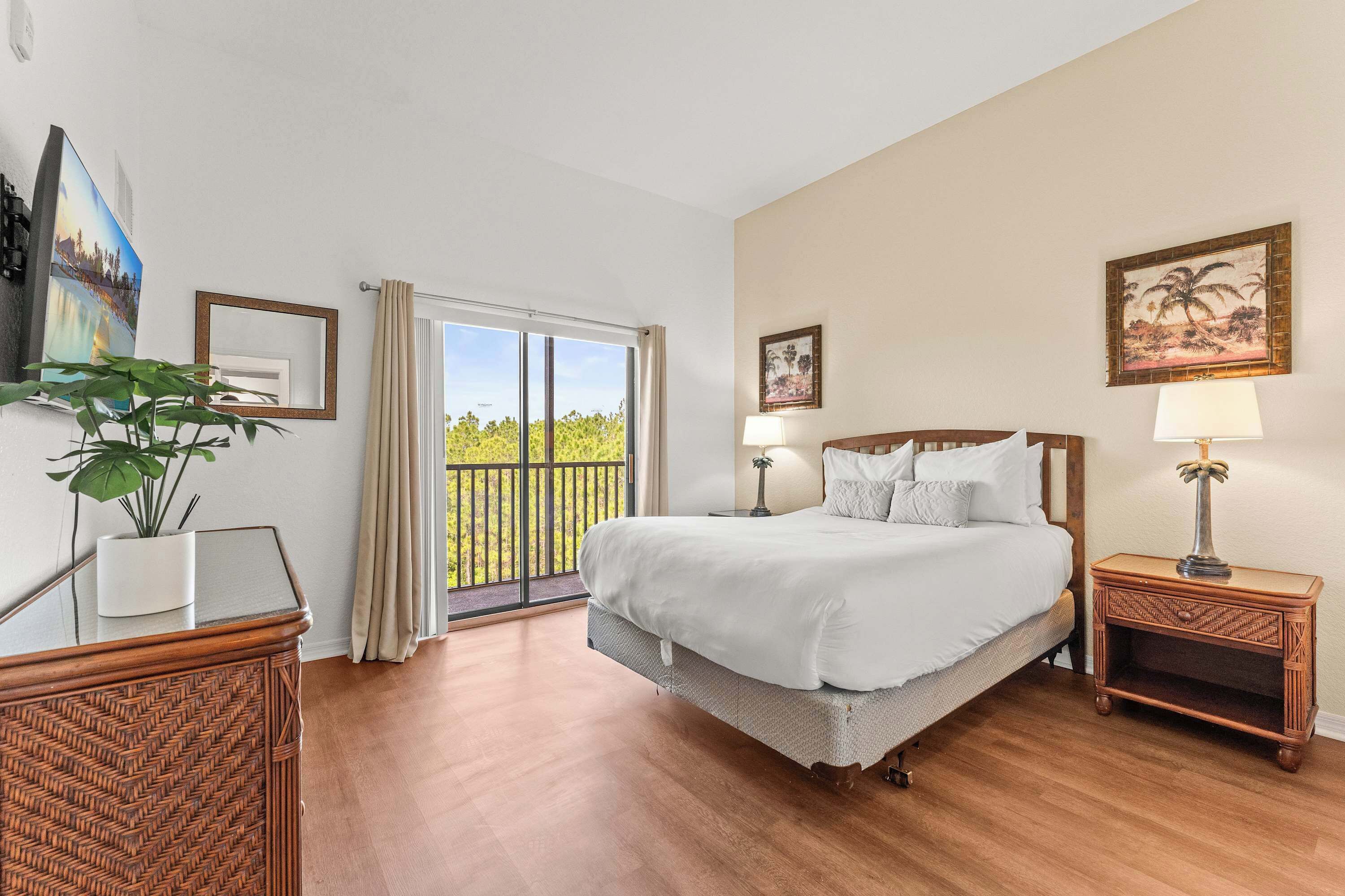 A bedroom with a large bed, a potted plant, a wooden nightstand, a painting, and a view of greenery through a window.