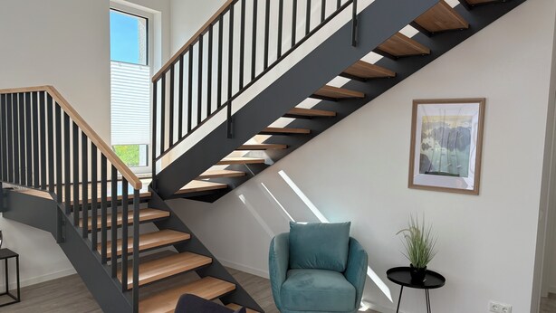 A modern staircase with wooden steps and black railings, a teal armchair, a small side table with a plant, and a framed picture on the wall. - Vacation home Wiesenblick (Spangenberg)