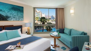 A hotel room with a bed, a sofa, a small table, and a view of a pool and buildings. - U Magic Palace (Eilat)