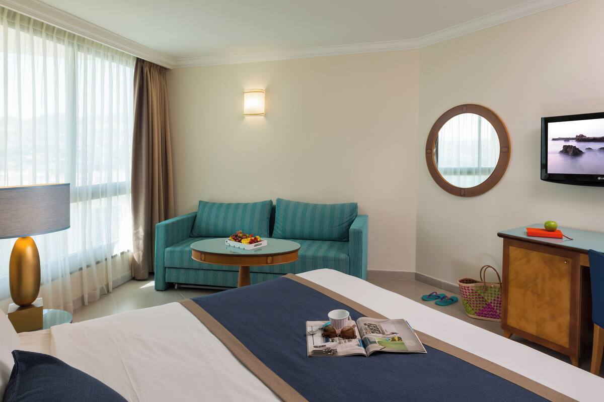 Une chambre d’hôtel avec un lit, un canapé, une petite table sur laquelle se trouve un panier de fruits, une télévision et une fenêtre avec des rideaux.