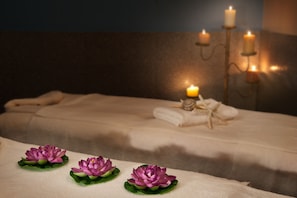 Una cama bien hecha con cubrecama blanco, una pequeña mesa con tres flores de loto moradas, y un candelabro de pared con velas.