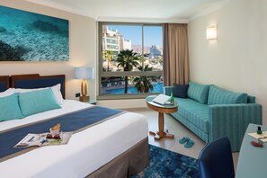 A hotel room with a bed, a sofa, a small table, and a view of a pool and buildings. - U Magic Palace (Eilat)