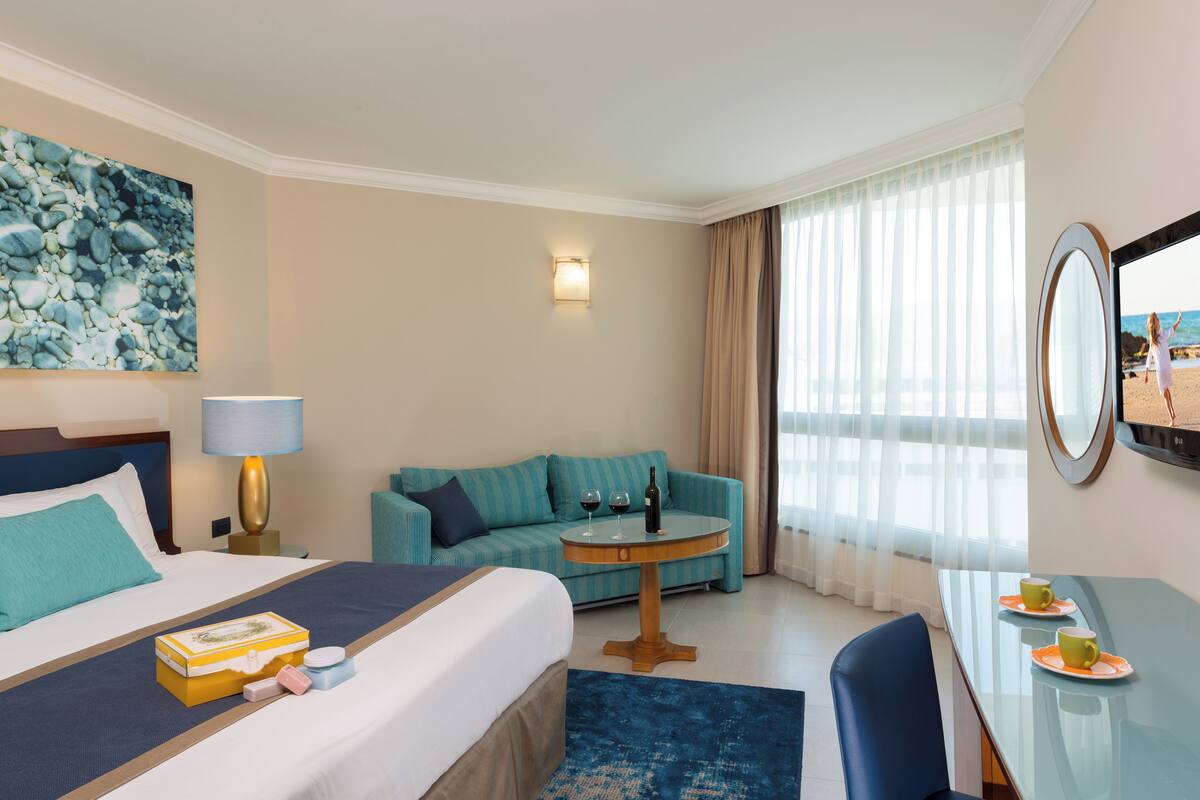 Une chambre d’hôtel avec un lit, un canapé, une table ronde avec une lampe, un bureau avec un miroir et un tableau au mur.