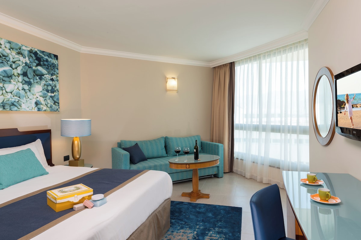 Una habitación de hotel con una cama, un sofá, una mesa redonda con una lámpara, un escritorio con un espejo y un cuadro en la pared.