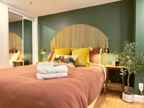 A bedroom with a bed, pillows, a bedside table, a lamp, and a potted plant.