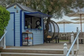 Un pequeño edificio azul con mostrador, posiblemente un bar de aperitivos, al lado de una gran estructura cilíndrica, probablemente un tobogán de agua, con asientos exteriores y sombrillas.