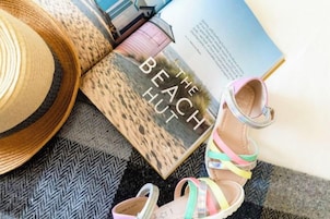 Un libro con el título 'The Beach Hut' y un par de sandalias coloridas sobre una alfombra estampada.