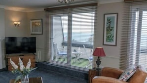 A living room with a TV, a sofa, a coffee table with flowers, and large windows with blinds. - Beachcroft Hotel, BW Signature Collection by Best Western (Felpham)