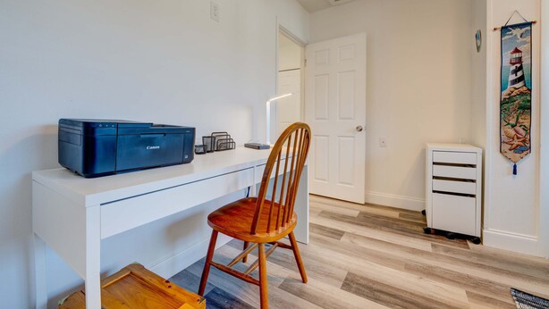 Una habitación con un escritorio blanco, una impresora Canon, una silla de madera, un armario blanco y un mueble blanco pequeño.