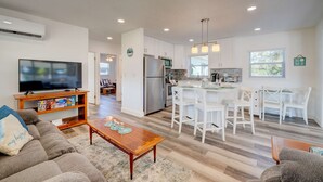 Una sala moderna con un televisor de pantalla plana, una mesa de centro de madera y un sofá gris. La cocina cuenta con muebles blancos, un refrigerador de acero inoxidable y un juego de comedor con sillas blancas.