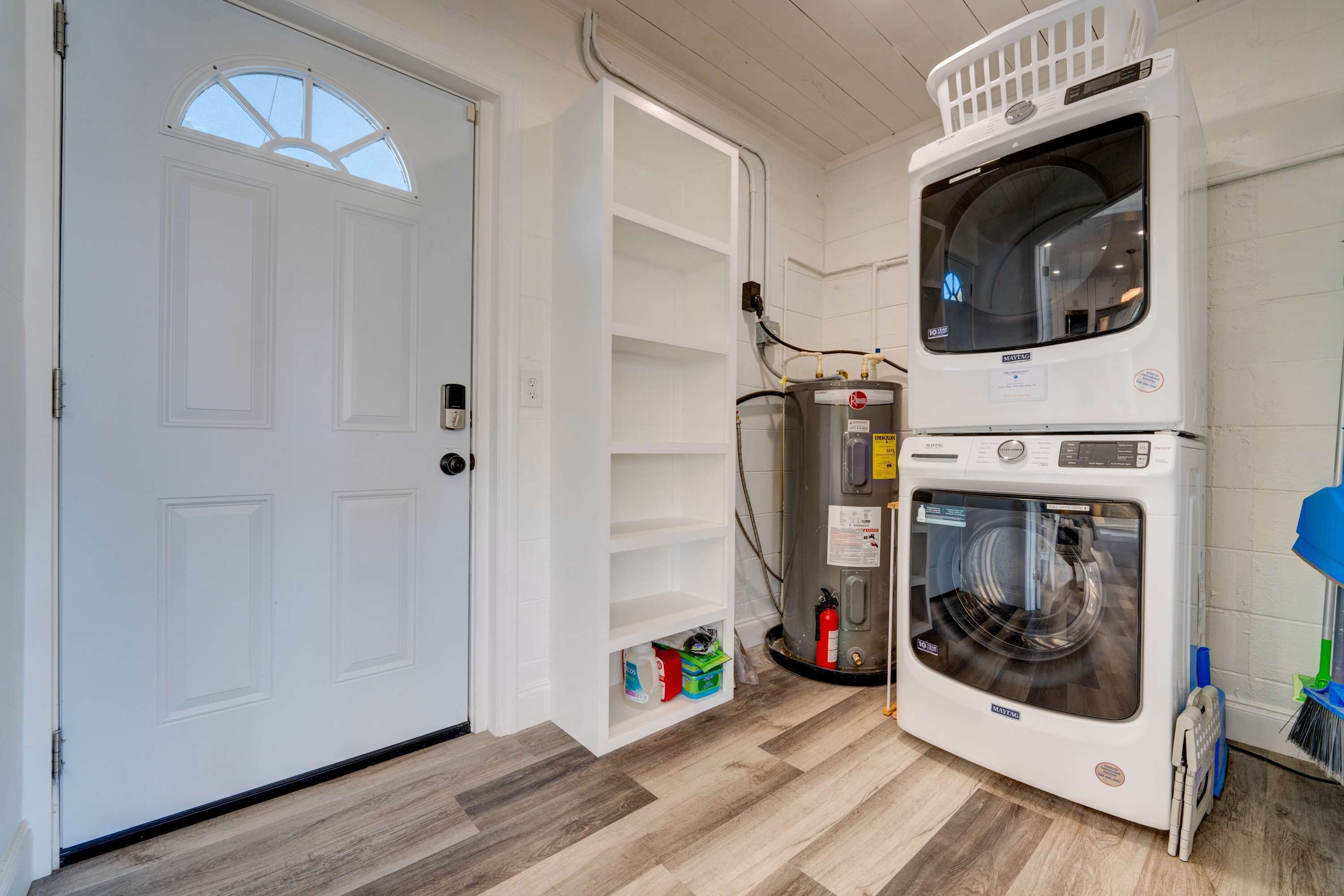 A laundry room with a stacked washer and dryer, a white door, and a shelf with cleaning supplies.