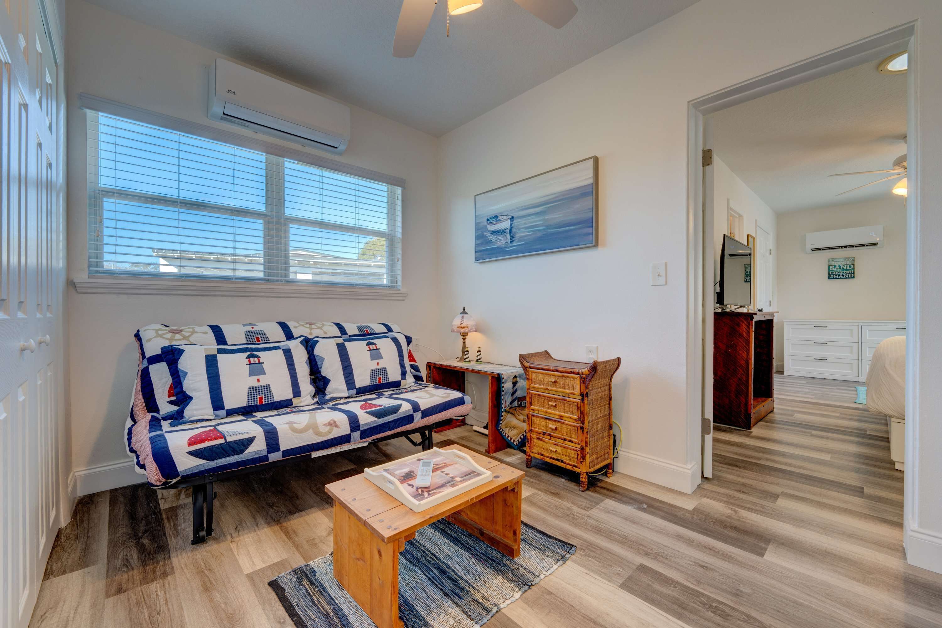 A living room with a sofa, a wooden coffee table, and a bench. There is a window with blinds, a painting on the wall, and a mirror above a dresser.