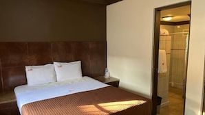 Une chambre d’hôtel avec un lit, une table de chevet, une salle de bain visible à travers une porte ouverte et un sol carrelé.