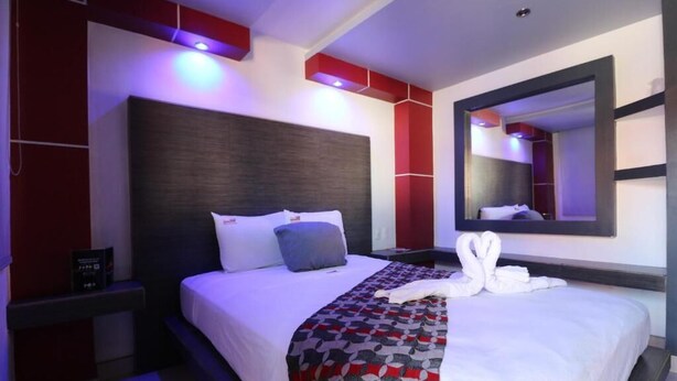 Une chambre d’hôtel moderne avec un grand lit, une table de chevet, un miroir et un mur d’accent rouge.