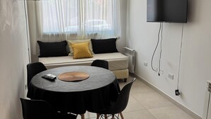 Ein modernes Zimmer mit einem Bett, einem an der Wand befestigten Fernseher, einem Esstisch mit Stühlen und einem großen Fenster mit transparenten Vorhängen.