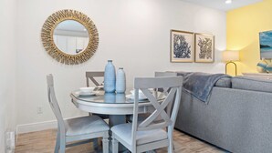 A dining area with a round table, chairs, and a wall-mounted mirror. There are framed artworks above the mirror and a sofa with a blanket in the background. - Capri Sunset (Kissimmee)