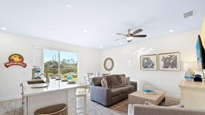 A modern living room with a grey sofa, a coffee table, and a dining area with a white table and chairs. A ceiling fan is visible, and there are framed artworks on the wall. - Capri Sunset (Kissimmee)