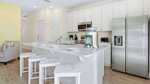 A modern kitchen with white cabinetry, a stainless steel refrigerator, a built-in microwave, and a central island with bar stools. - Capri Sunset (Kissimmee)