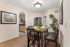 Un coin repas avec une table en bois dressée pour quatre personnes, entourée de chaises en bois foncé. Un tableau encadré est accroché au mur et une plante en pot se trouve à droite.