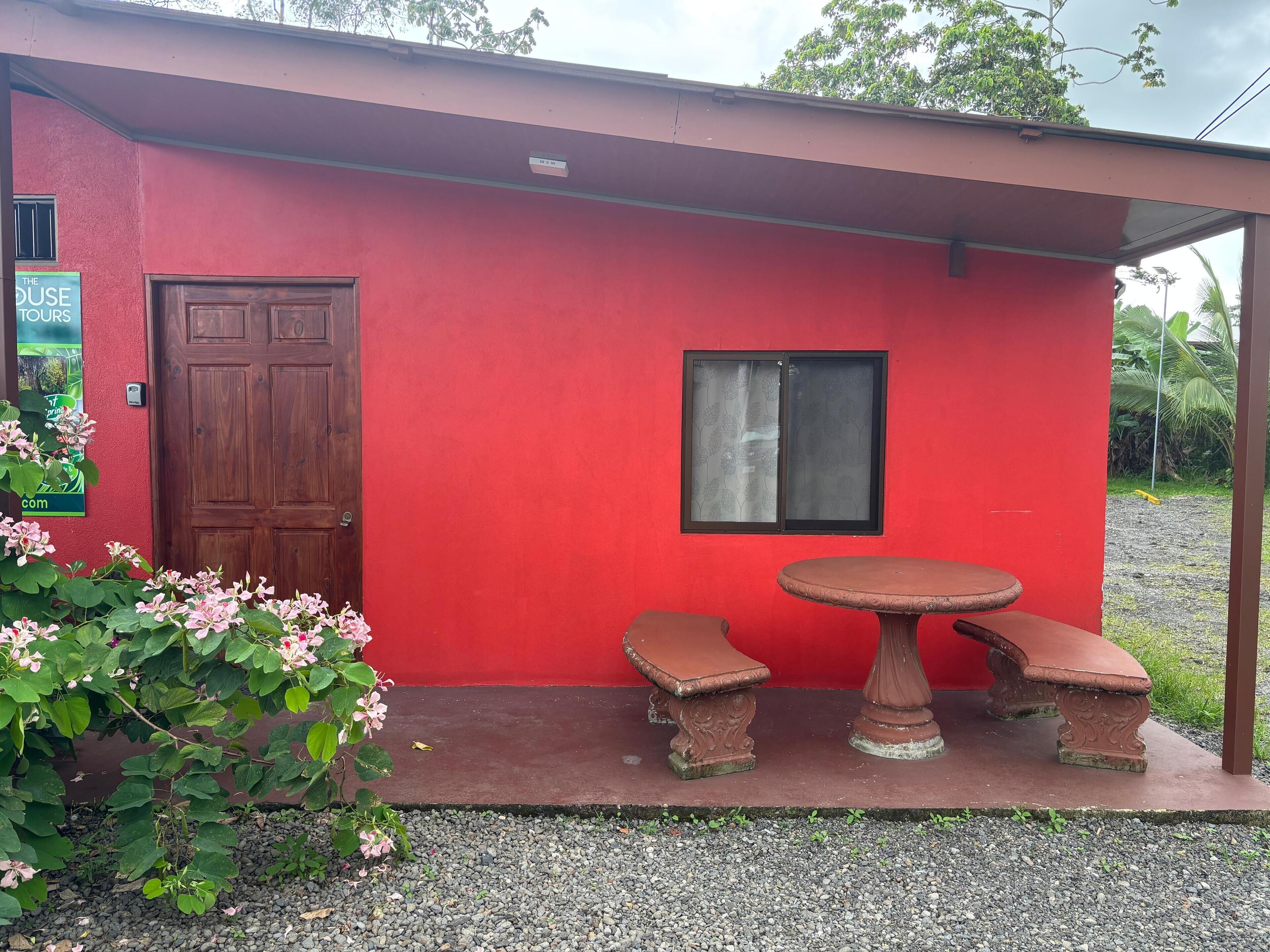 Un bâtiment rouge avec une porte en bois, une fenêtre et un petit espace extérieur aménagé avec une table et des bancs.