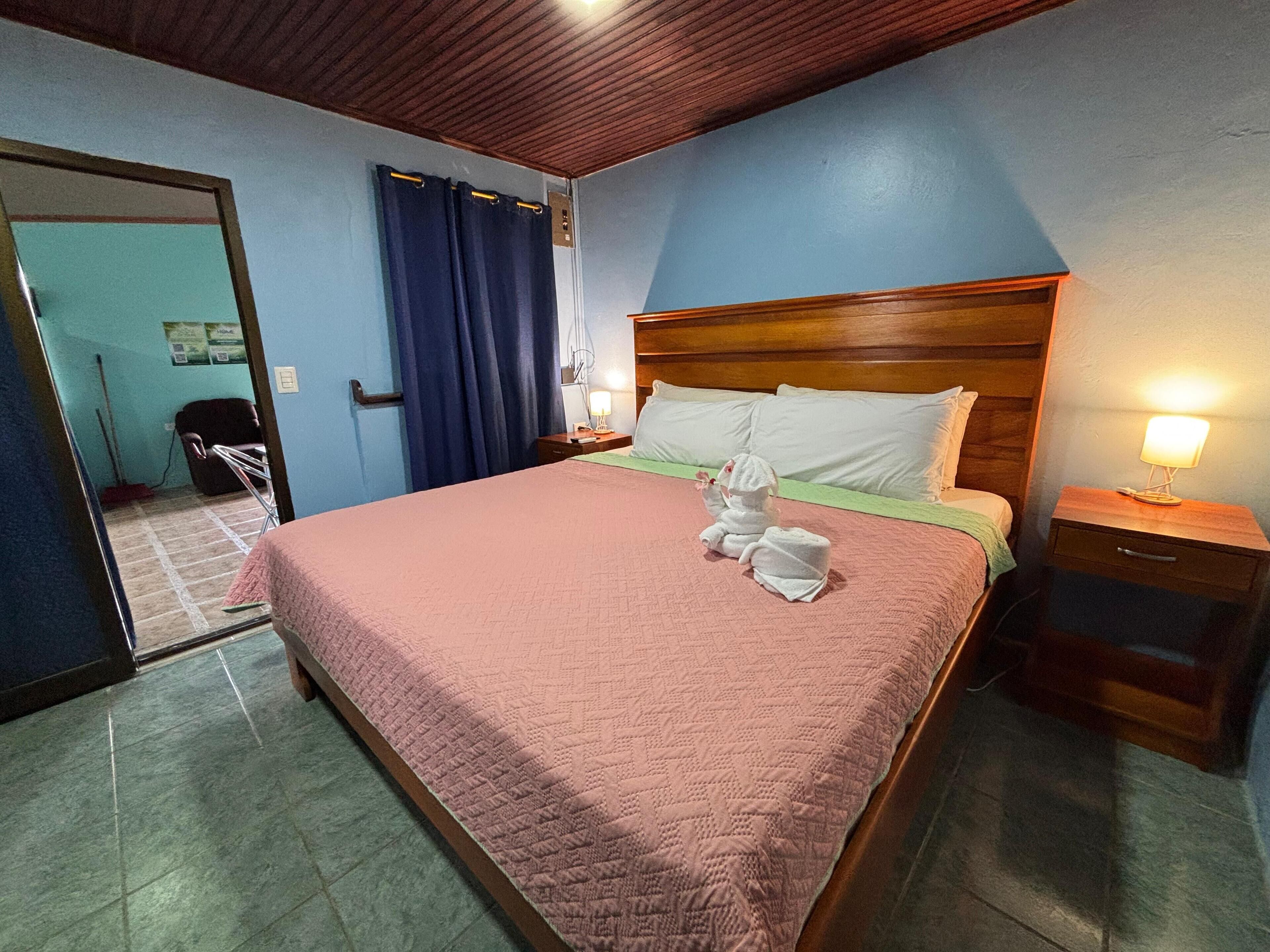 Hotelkamer met een houten hoofdeinde, een roze gewatteerde spreideken, een nachtkastje met een lamp en uitzicht op de gang.