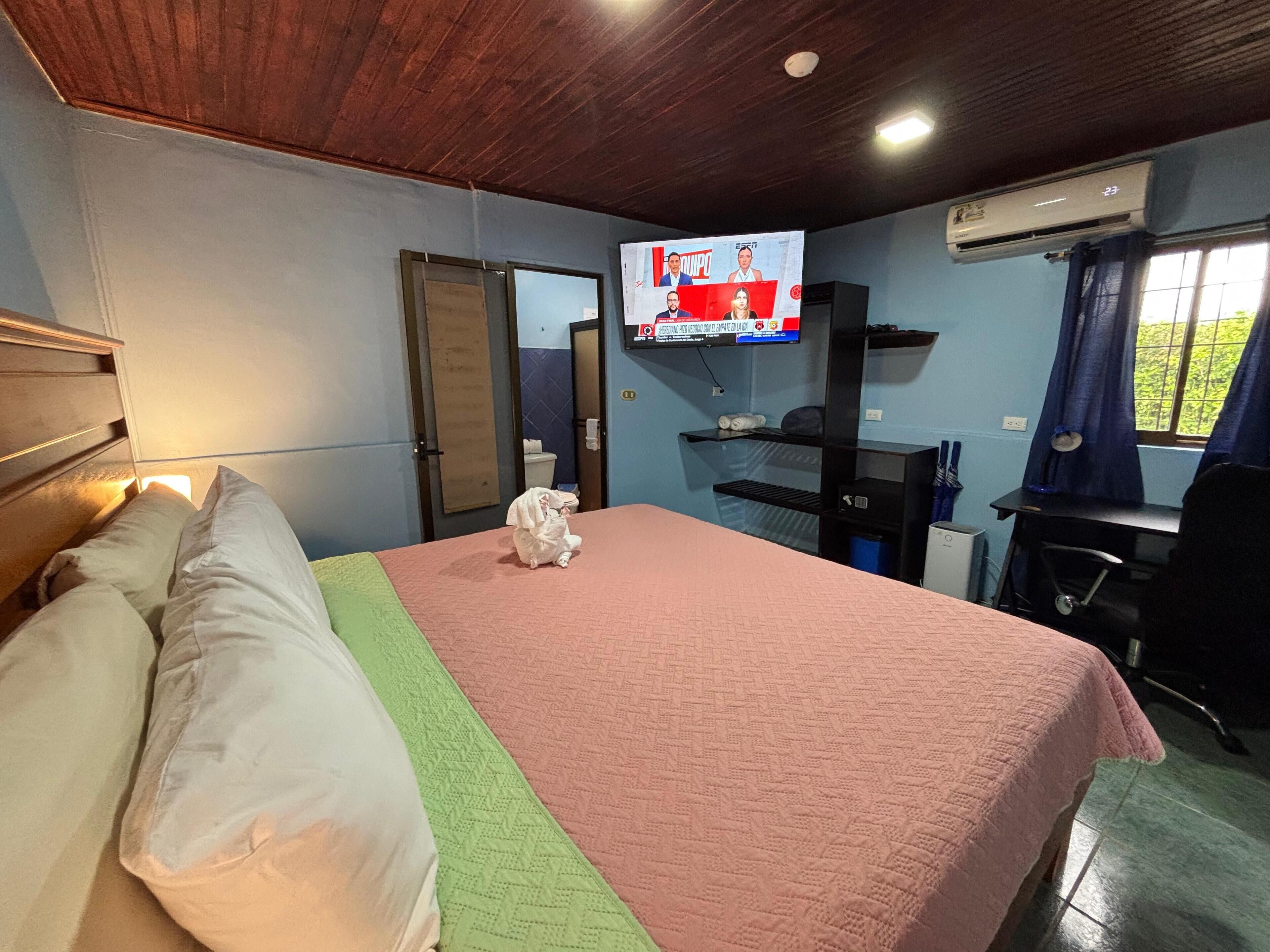 Habitación de hotel con cama, televisor montado en la pared, escritorio con silla y ventana con cortinas.