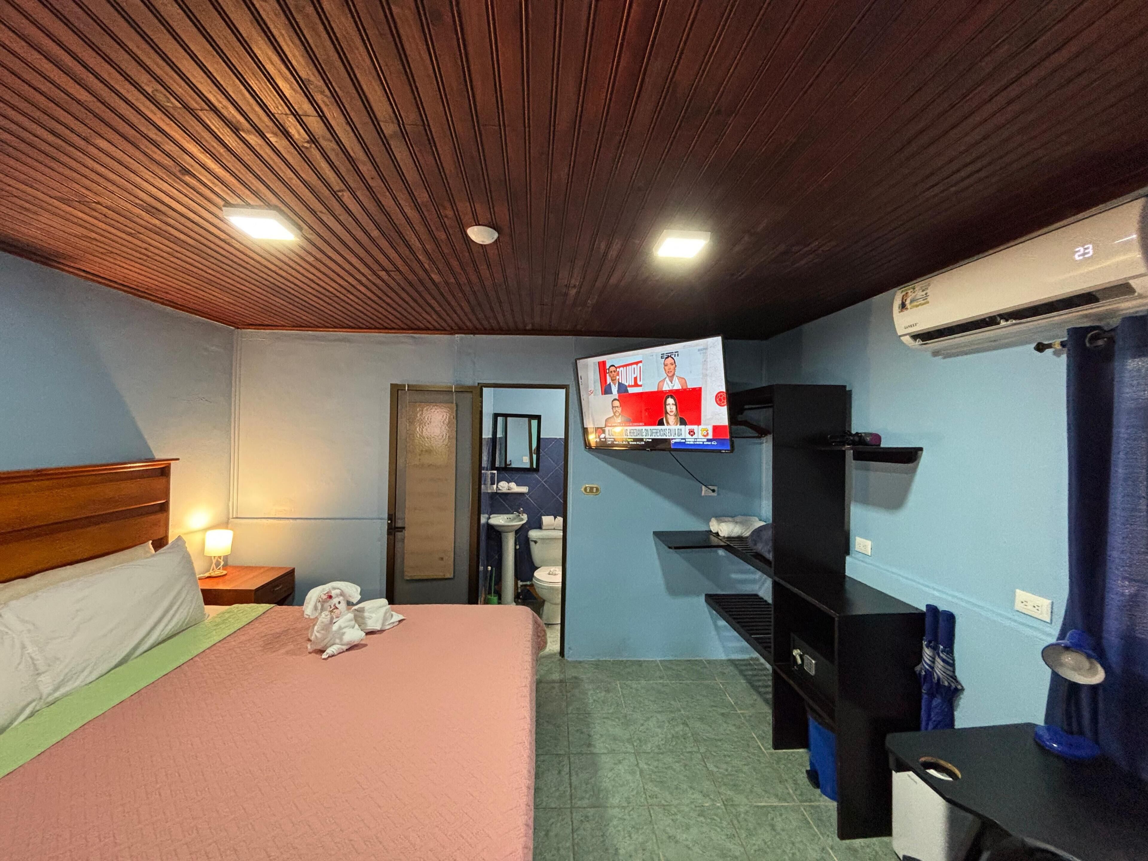 Ein Hotelzimmer mit Holzboden, einem Bett mit pinkfarbener Tagesdecke, einem an der Wand befestigten Fernseher, einem Waschbecken und einem blauen Vorhang.
