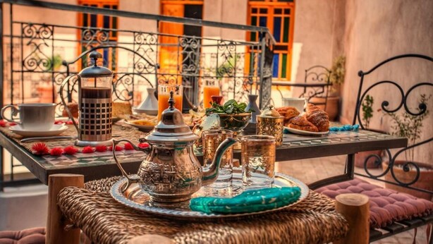 Ett bord dukat med ett traditionellt marockanskt teservis, inklusive en kaffekanna, tekanna, koppar och glas, tillsammans med ett brickbord i rotting och kuddar.