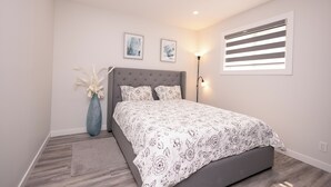 A bedroom with a bed, a nightstand, a vase, and a window with blinds. - Cozy Cove Extension (Winnipeg)