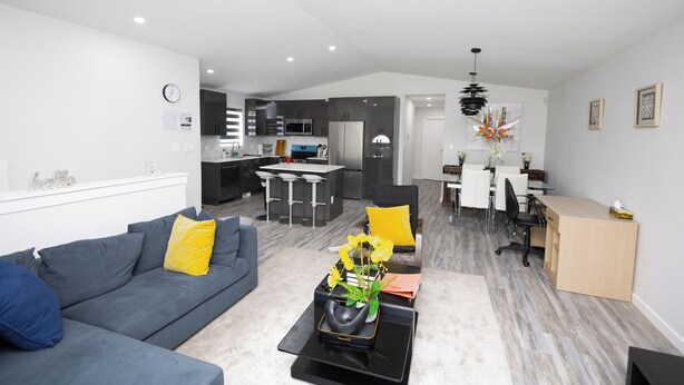 A modern living room with a gray sofa, a coffee table with a vase of flowers, and a dining area with a kitchen in the background. - Cozy Cove Extension (Winnipeg)