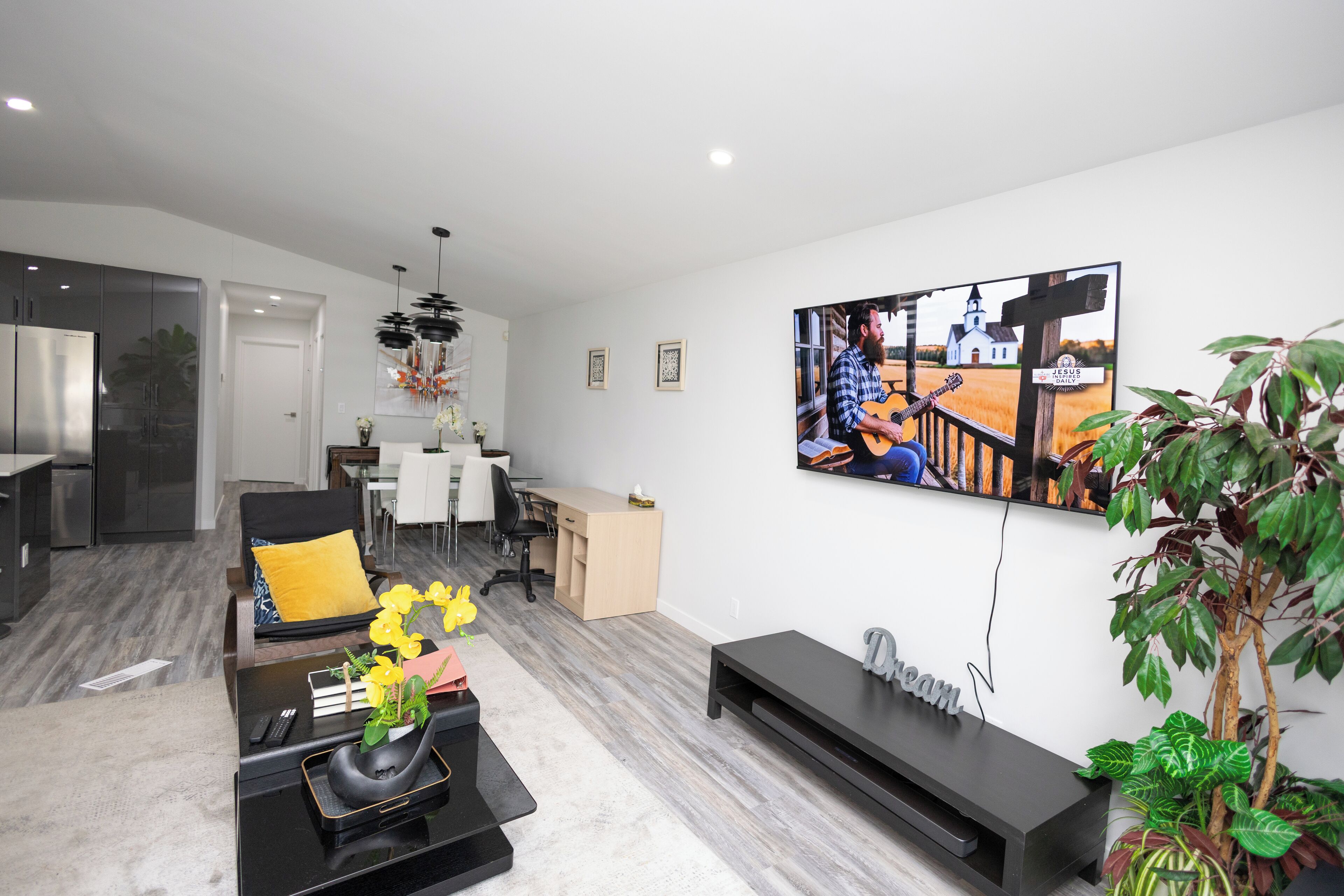 Een moderne woonkamer met een flatscreen televisie, een bijzettafeltje met bloemen, een bank met een geel kussen, en een eethoek met een witte tafel en stoelen.