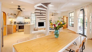 A modern kitchen with a central island, a dining area with a wooden table, and a living space with a TV and plants.
