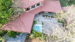 A multi-level house with a red tiled roof, a swimming pool, and lush greenery surrounding the property.