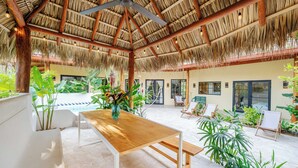 A spacious indoor area with a thatched roof, wooden tables, and chairs, surrounded by lush greenery.