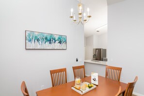 A dining area with a wooden table, four chairs, a chandelier, and a painting on the wall. - Celebrate at Club Cortile (Kissimmee)