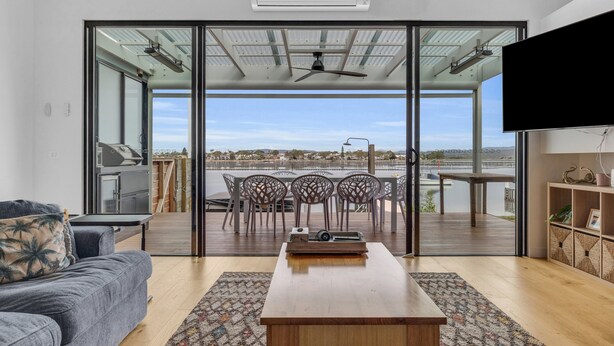 A modern living room with a sofa, coffee table, and a TV. It features a large glass door leading to an outdoor deck with a view of the water and cityscape. - Lake Front: 3 Bedroom: Spa (Merimbula)