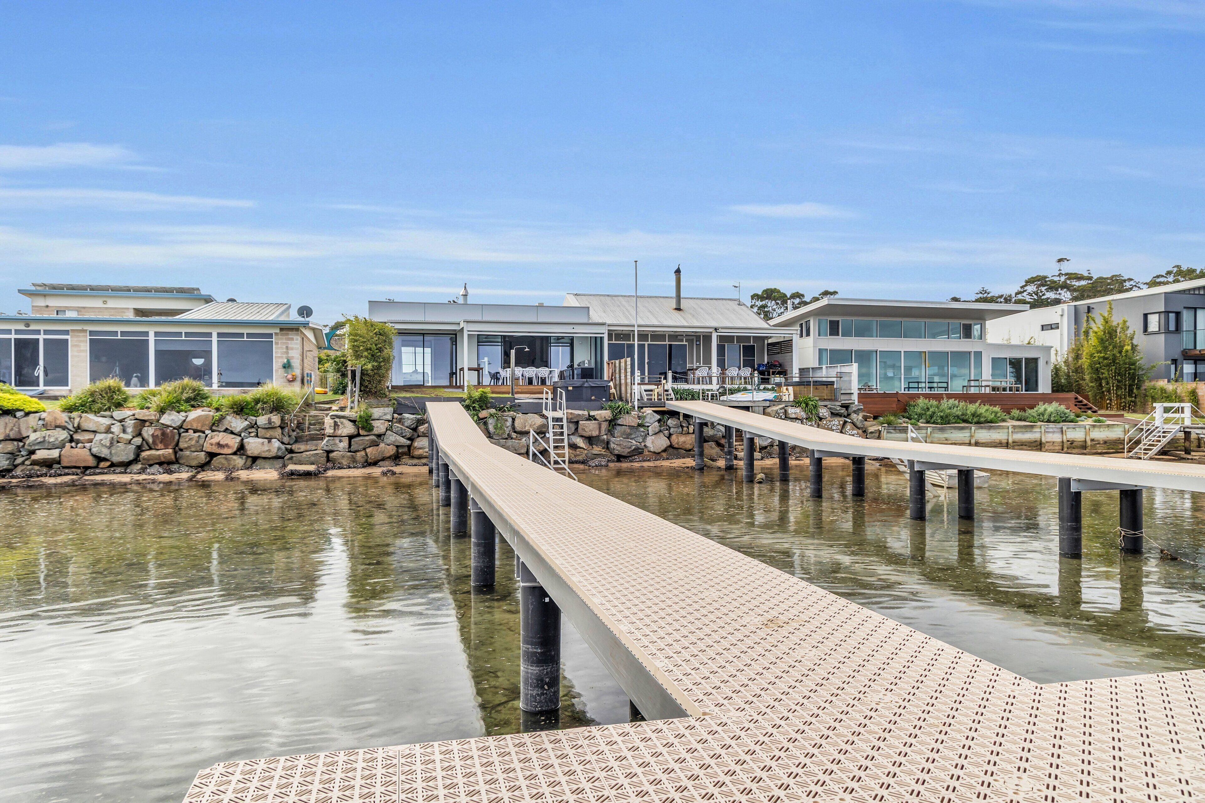 A waterfront property with a dock extending into a calm body of water, modern buildings with large windows, and a stone wall along the water's edge.