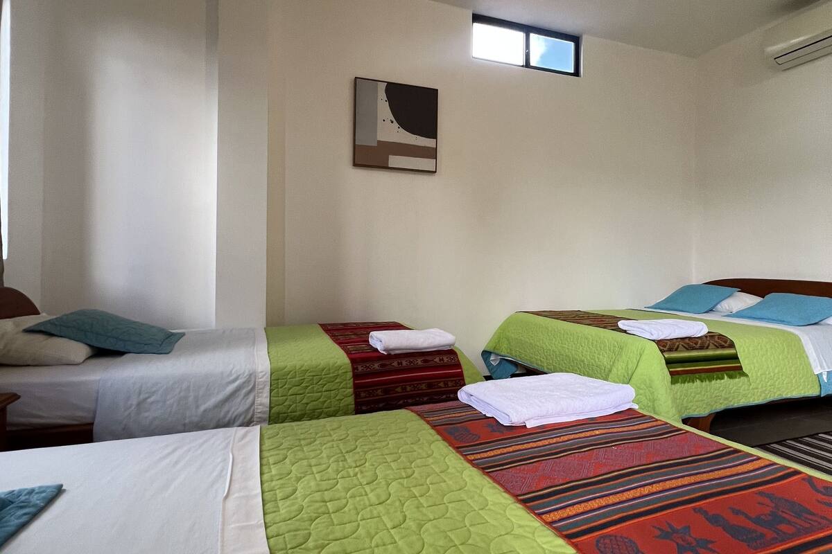 Habitación de hotel con tres camas, cada una con ropa de cama de diferente color, un ventanal pequeño y una obra de arte colgada en la pared.