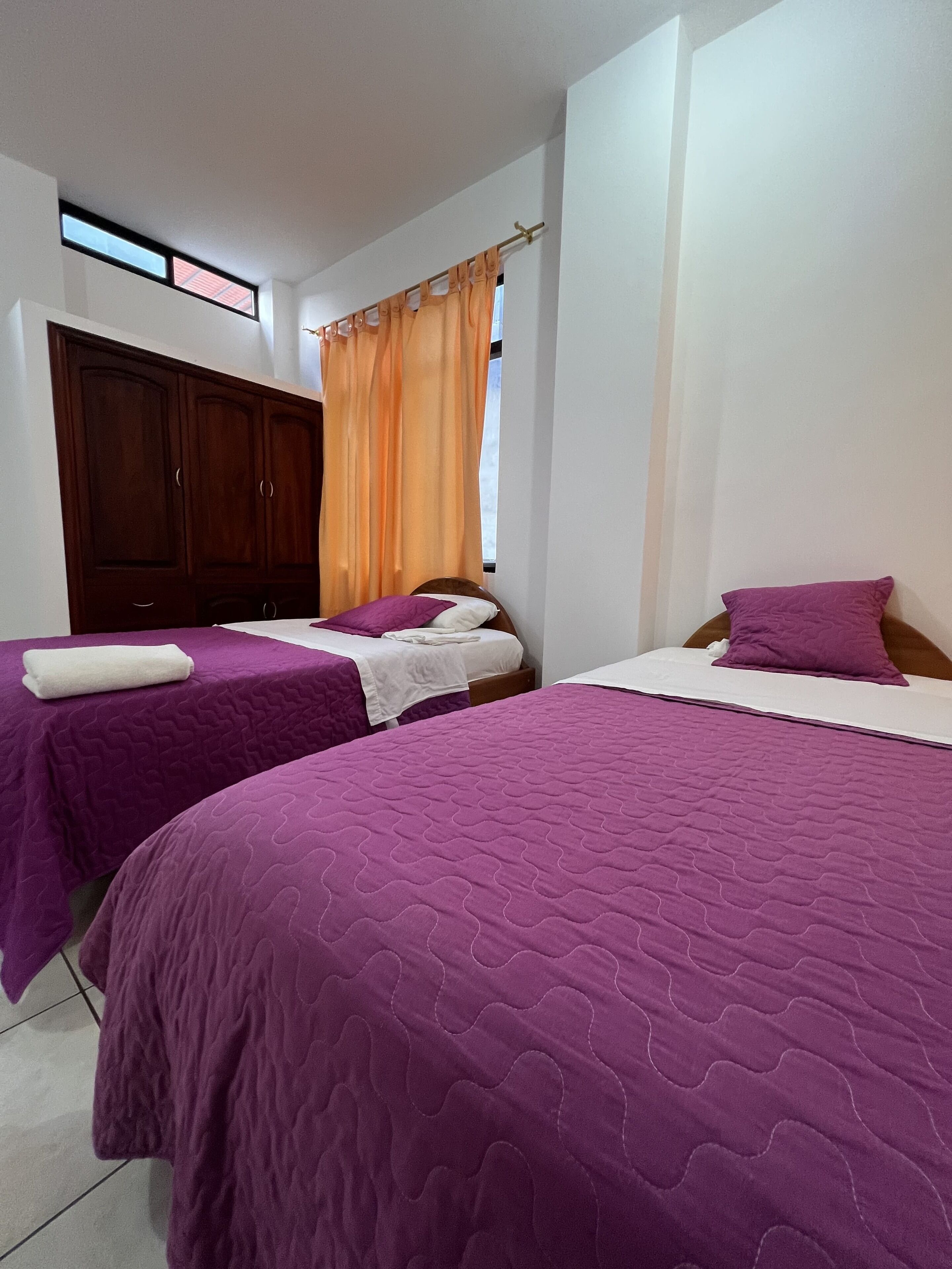A bedroom with two beds, each with a purple bedspread, a wooden wardrobe, and a window with orange curtains.