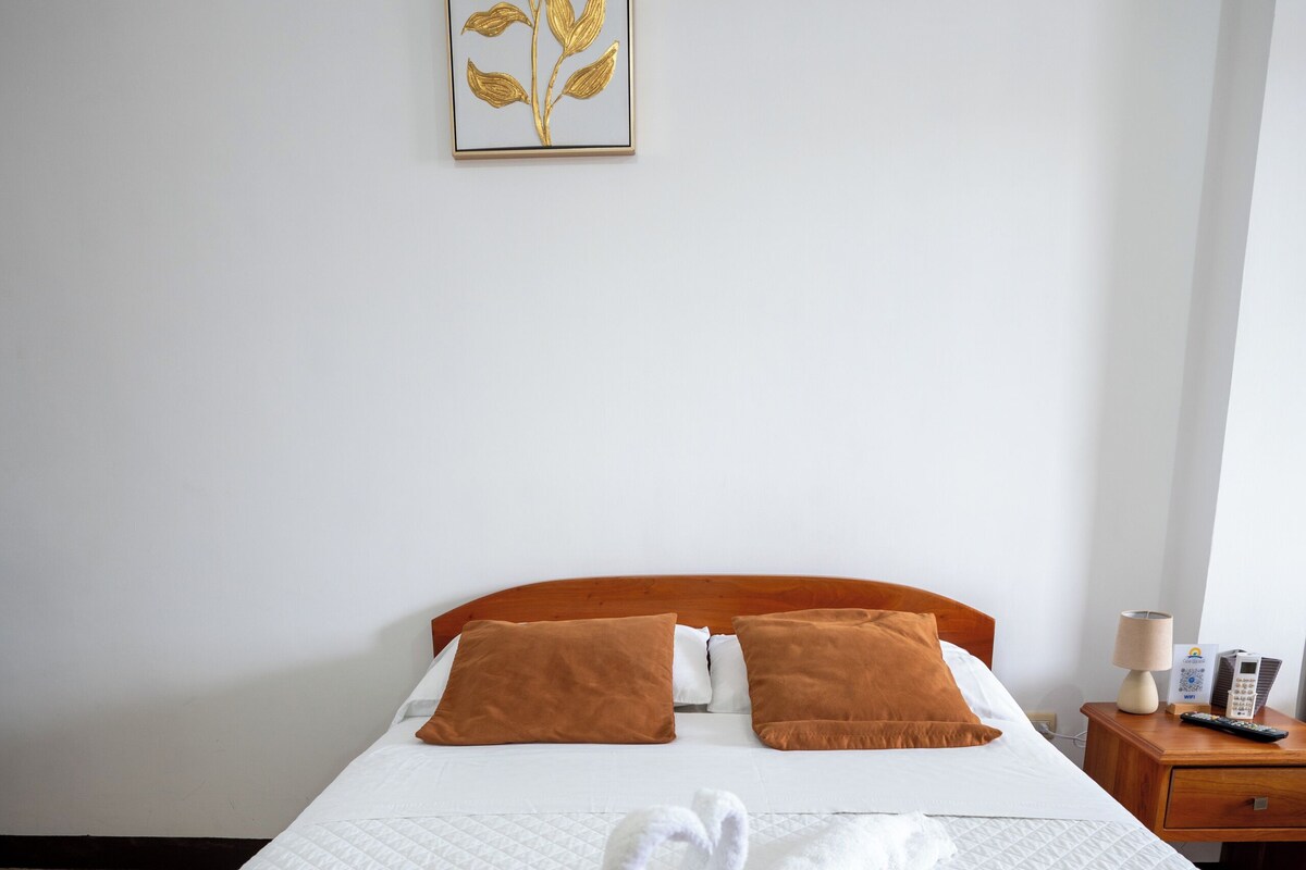 Una cama bien hecha con cabecera marrón, ropa de cama blanca y una obra de arte con forma de hoja dorada en la pared.