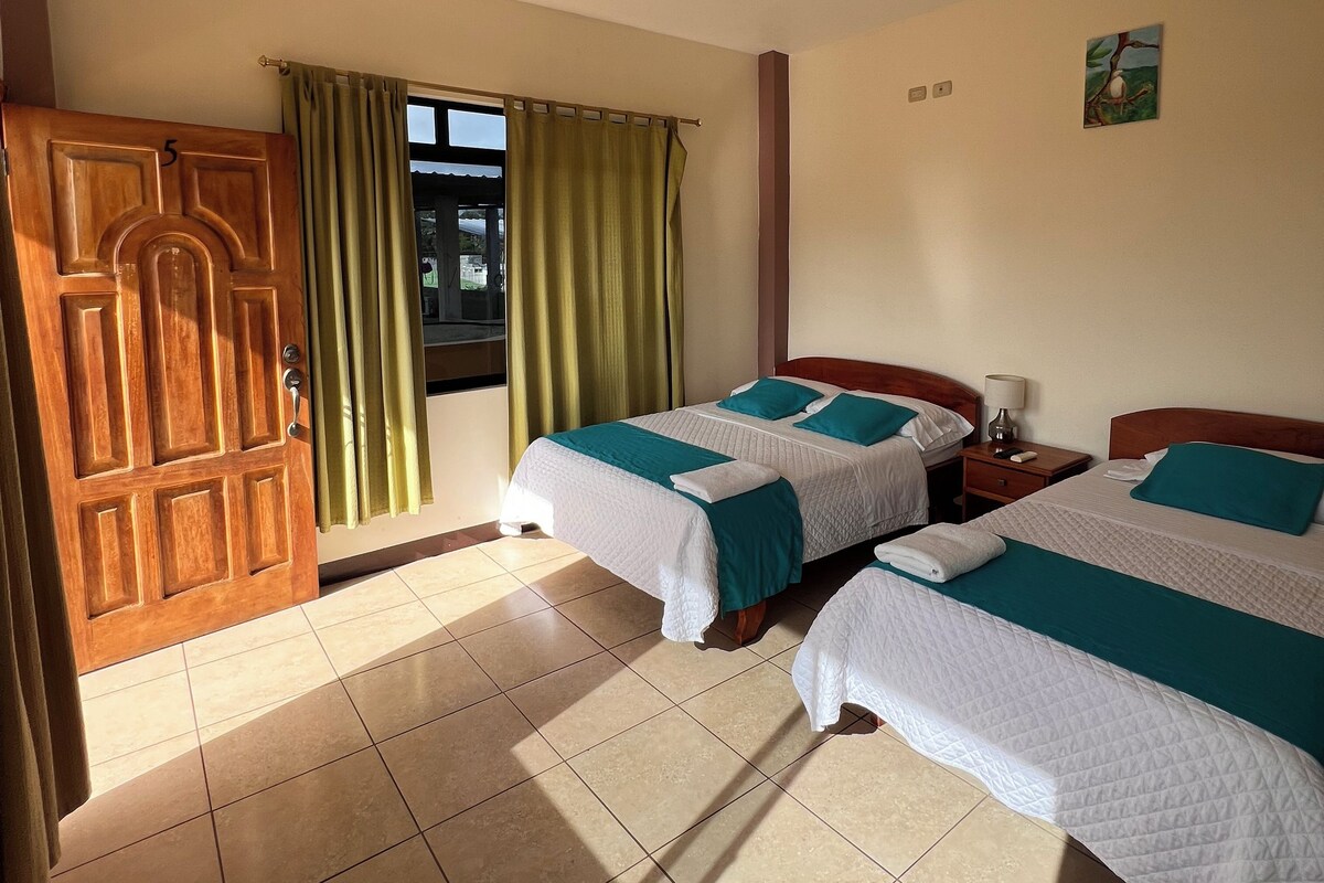 A hotel room with two beds, a wooden door, a window with curtains, a nightstand, and a wall-mounted light fixture.