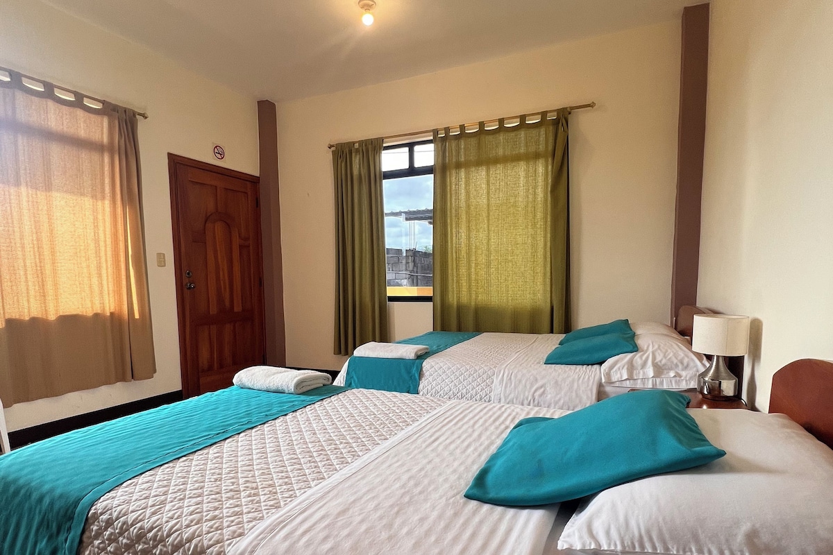 A hotel room with two beds, a window with curtains, a door, and a ceiling light.