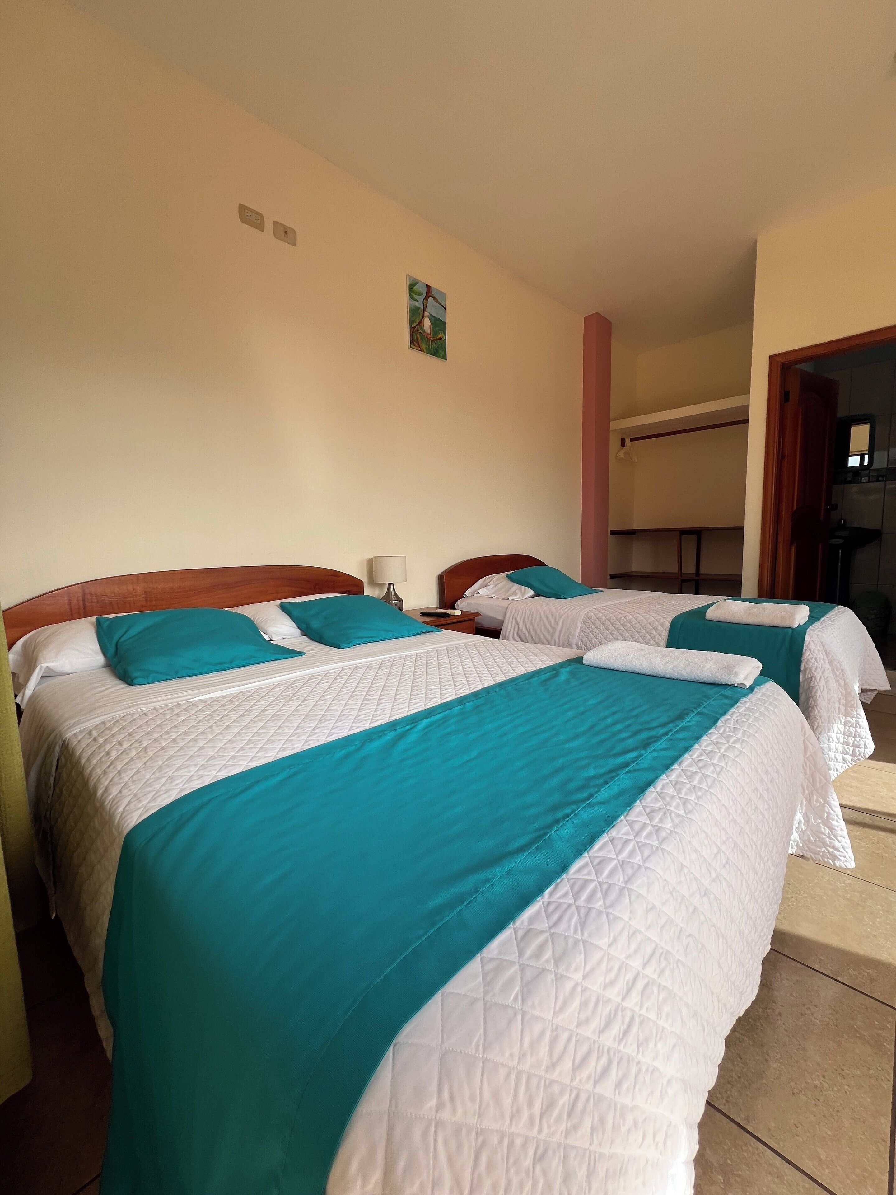 Una habitación de hotel con dos camas, cada una con una colcha verde azulada y blanca, una mesita de noche pequeña con una lámpara, y un cuadro colgado en la pared.
