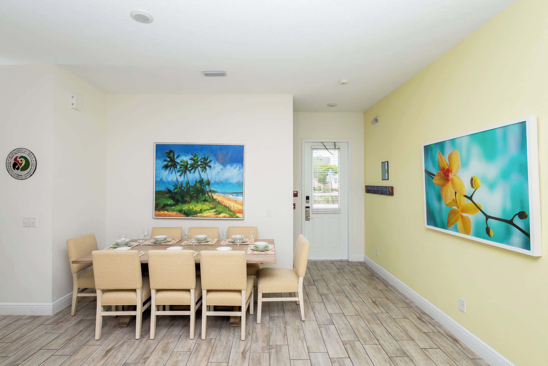 Un coin repas avec une table dressée pour six personnes, entourée de chaises de couleur beige, et un tableau représentant des palmiers et une plage accroché au mur.