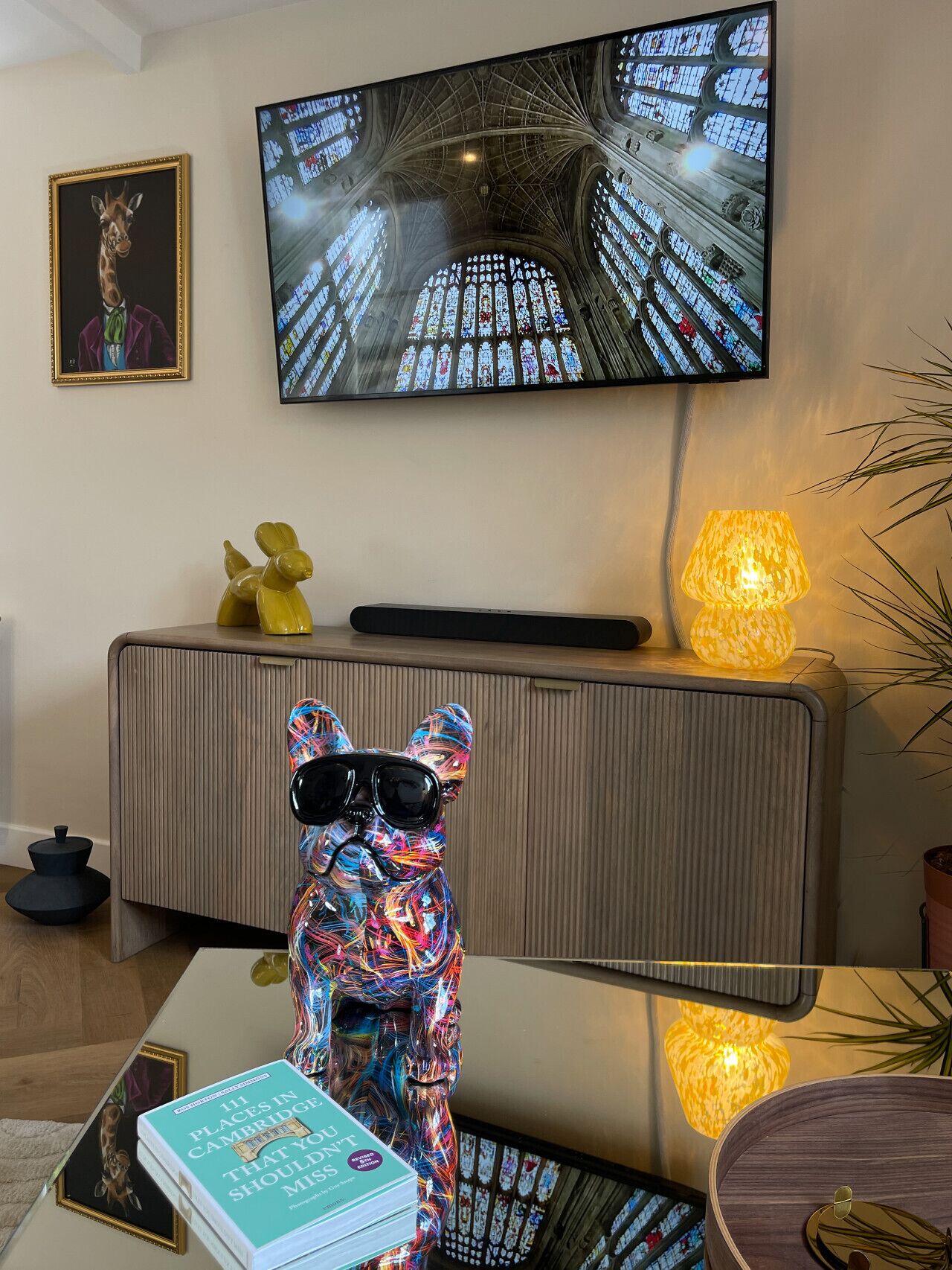 A modern living room with a television displaying a cathedral interior, a decorative dog sculpture, and a book titled "111 Places in Cambridge You Don't Should Miss".