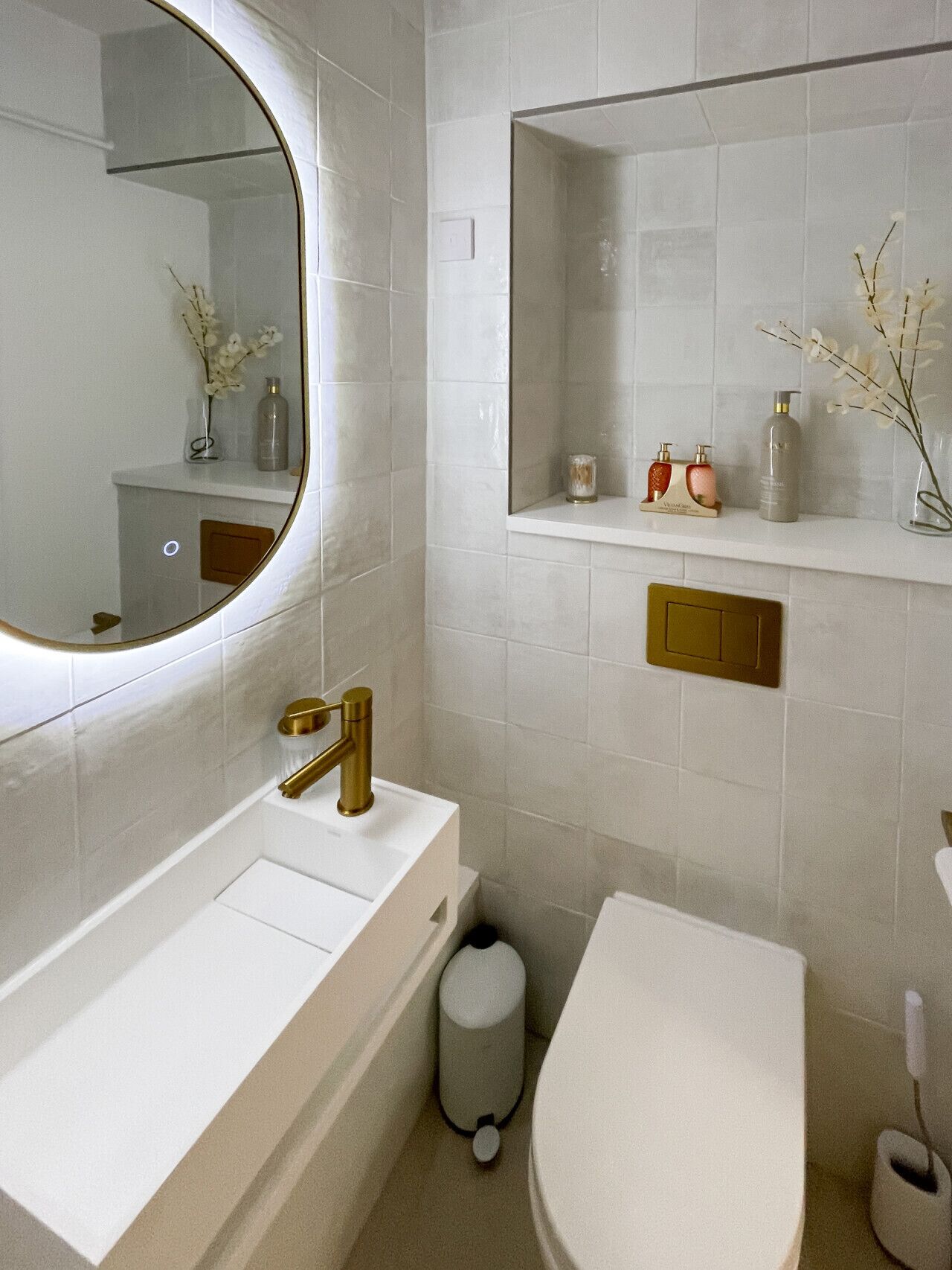 A modern bathroom with a white sink, gold faucet, and a round mirror. There is a toilet and a trash can.