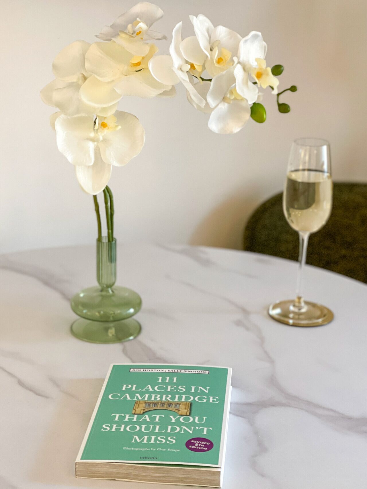 En bok med titeln ”111 platser i Cambridge som du inte får missa” ligger på ett bord av marmor, tillsammans med en vas med vita orkidéer och ett glas vitt vin.